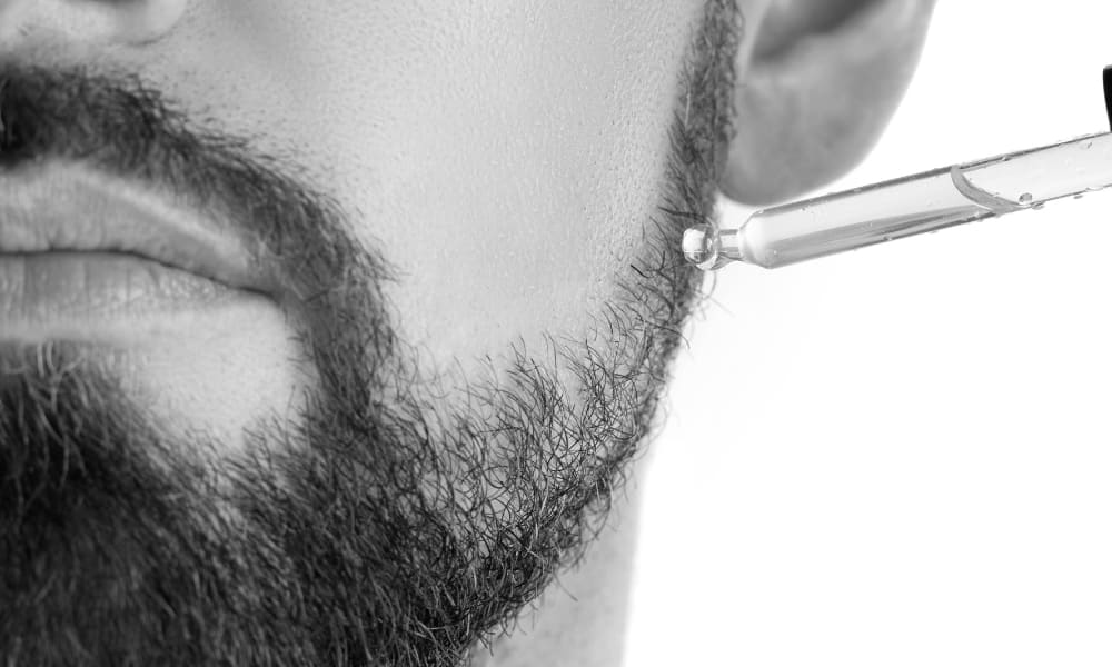 Close-up of man applying beard oil with a dropper to hydrate facial hair and skin, supporting beard maintenance and healthy growth.