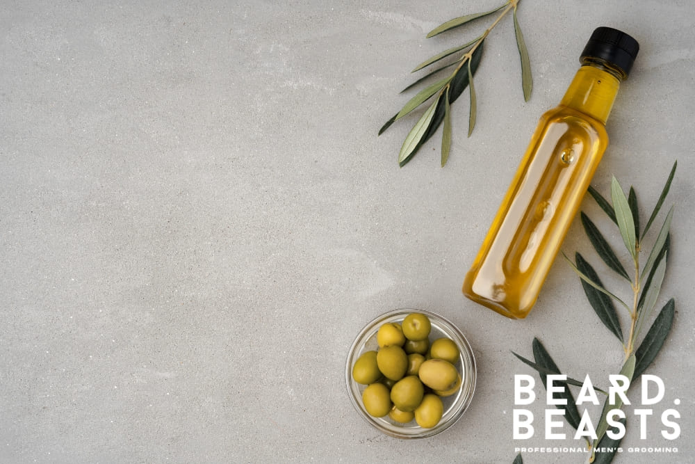 A glass bottle of olive oil and a small bowl of fresh green olives placed on a light, textured surface, surrounded by olive branches. This image highlights the natural ingredients used in olive oil for beard care, showcasing its purity and suitability for nourishing and softening facial hair