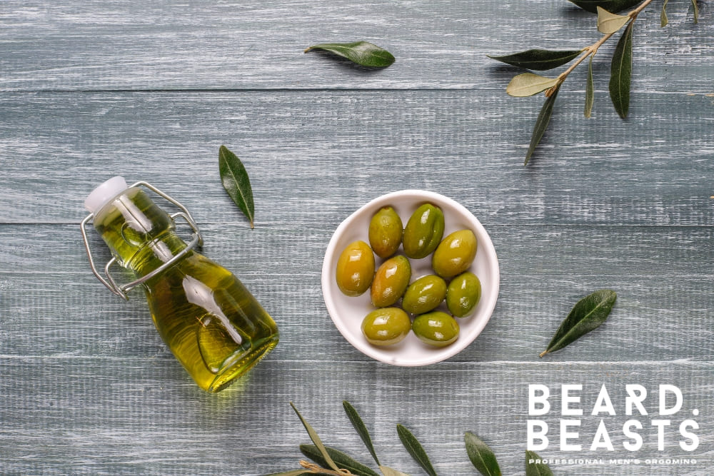 A glass bottle of olive oil and a white bowl filled with fresh green olives placed on a rustic wooden table, surrounded by olive leaves. Perfect representation of natural ingredients for olive oil in beard care.