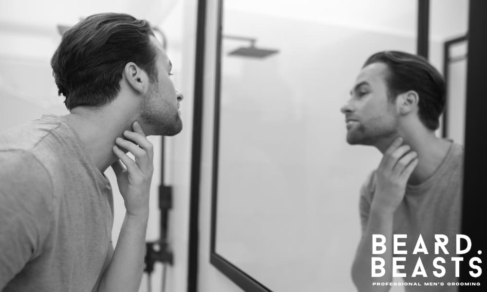 Man examining early stubble growth in the mirror, assessing beard density and neckline placement for a well-shaped designer stubble style.