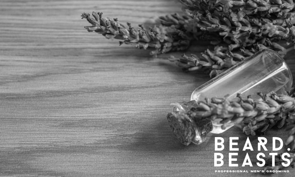 Small glass bottle of lavender essential oil surrounded by fresh lavender sprigs on a wooden surface—used in natural beard care routines.