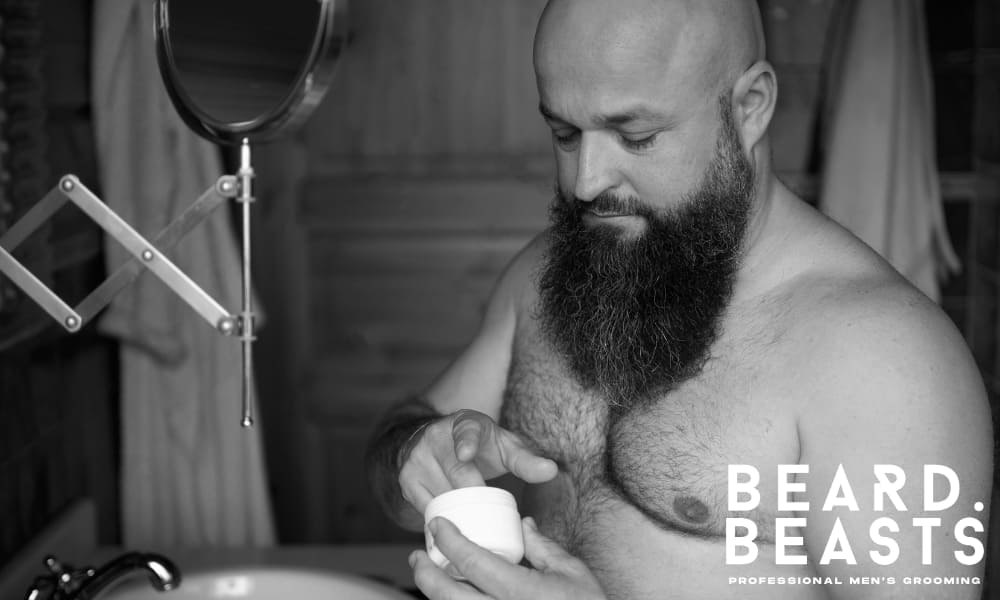 Bald man with a full beard applying beard balm in front of a bathroom mirror as part of his grooming routine