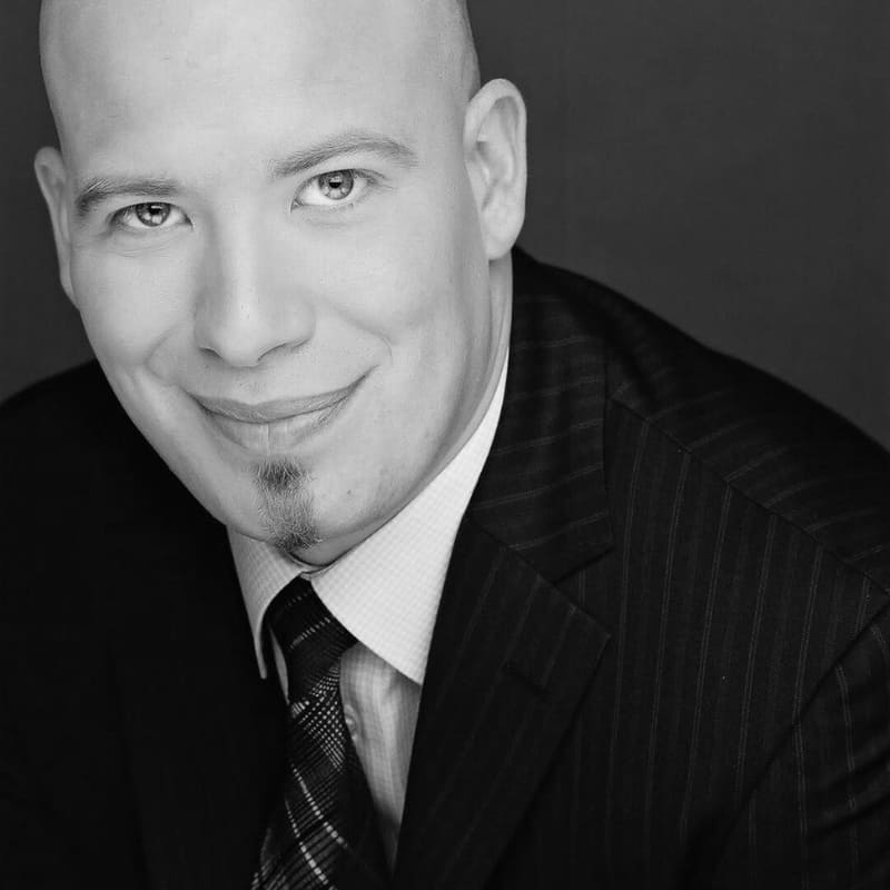 Man wearing a dark pinstripe suit and tie, smiling slightly with a neatly groomed patch of facial hair below the lower lip, against a red background.