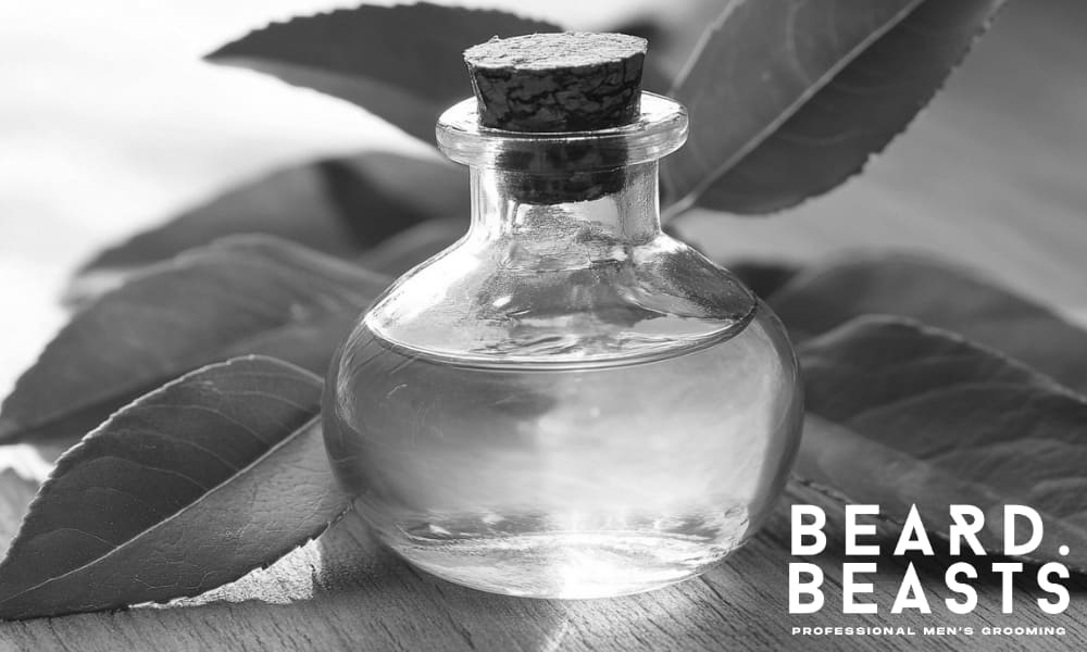 Glass bottle of tea tree essential oil with green leaves on a wooden surface—used to cleanse and support healthy beard growth.