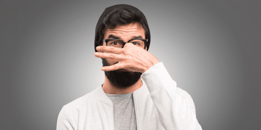 Young Man Wearing Glasses and a Beanie, Holding His Nose Due to Strong Beard Odor - Learn Expert Tips to Prevent and Eliminate Bad Beard Smells and Maintain Freshness