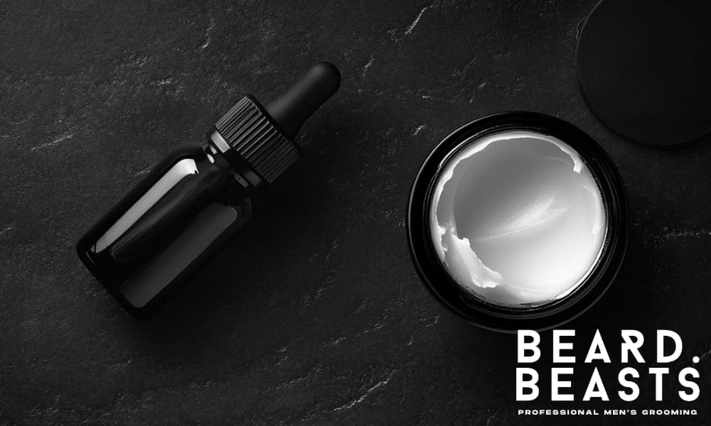 Flat lay of beard oil bottle with dropper and open tin of beard butter on a dark background