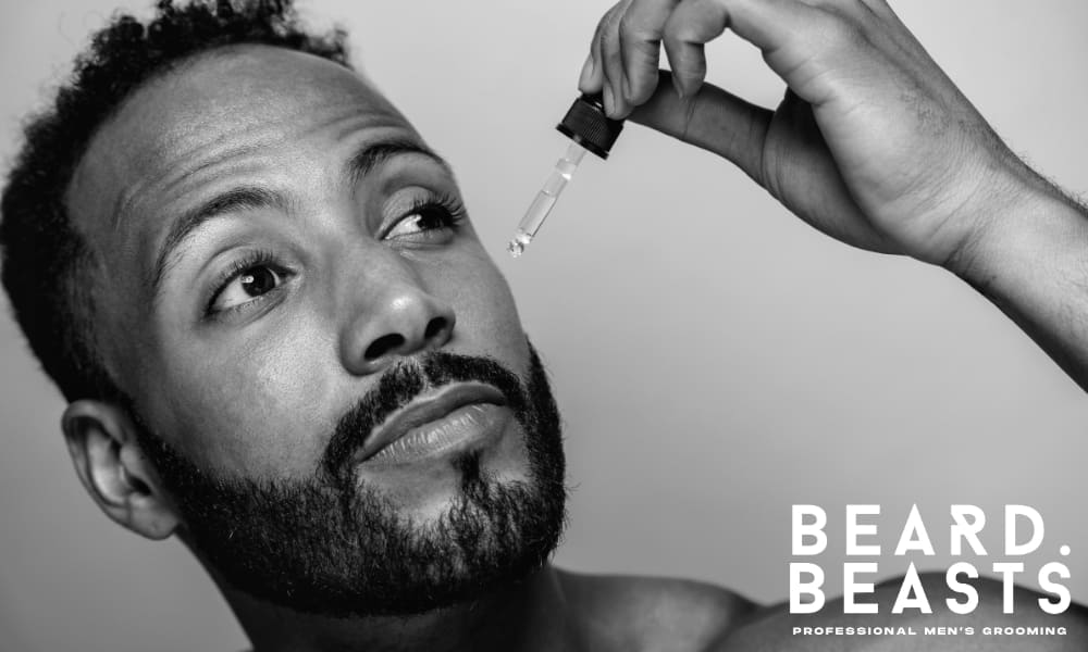 Close-up of a bearded man applying beard oil with a dropper, highlighting skin hydration and beard care routine.