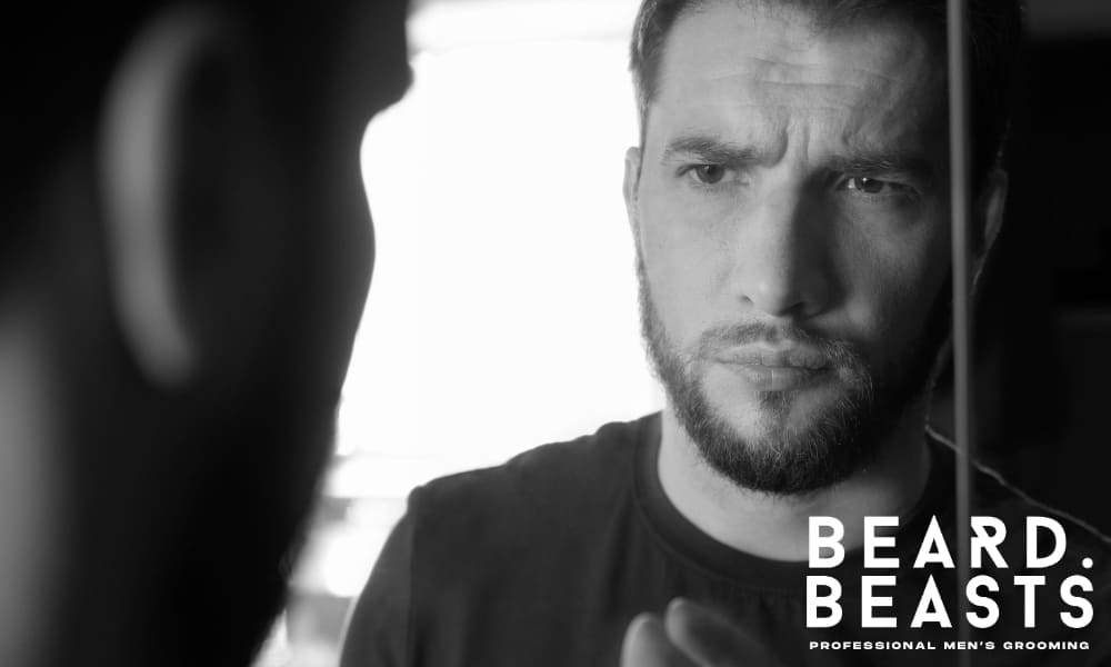 Man inspecting his facial hair in the mirror while learning how to grow a full beard step by step