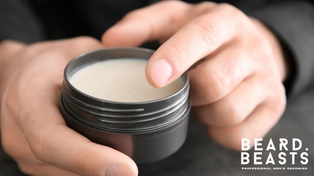 Close-up of hands holding a tin of beard balm, with one finger gently scooping the creamy product. The image highlights the smooth texture and practical use of beard balm for conditioning and grooming facial hair.