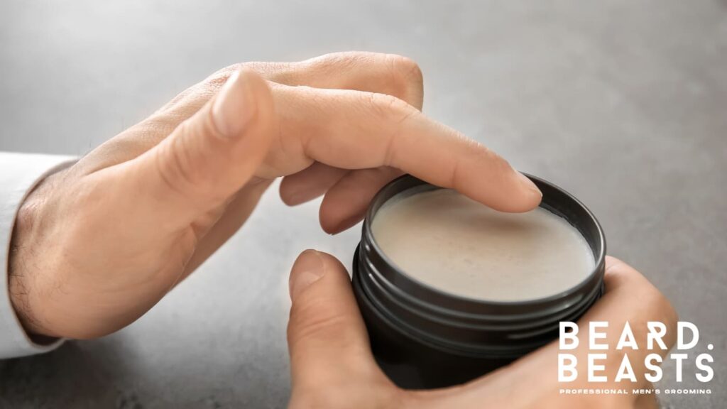Close-up of hands using a tin of beard balm, showcasing its creamy texture as one finger scoops the product. The image highlights the difference in application and consistency between mustache wax vs beard balm, emphasizing the balm's moisturizing and conditioning properties.