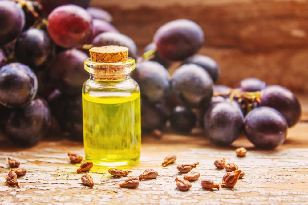 Small glass bottle filled with light golden oil, surrounded by scattered seeds and a bunch of fresh purple grapes in the background, set on a rustic wooden surface.