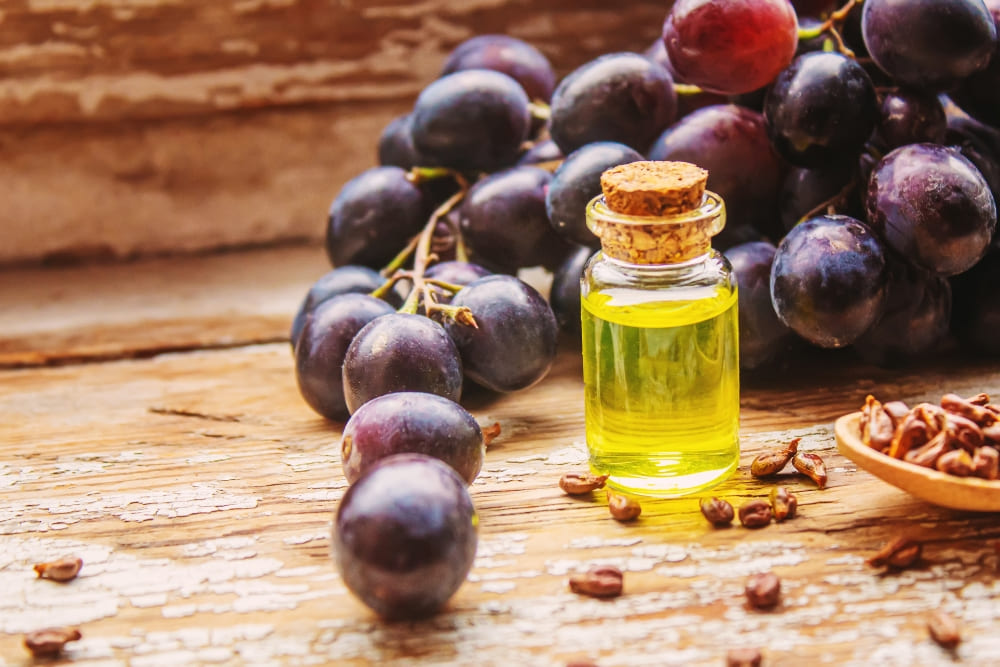 Small glass bottle of golden grapeseed oil for beard care, surrounded by fresh purple grapes and scattered seeds, placed on a rustic wooden surface with natural light enhancing the rich colors.