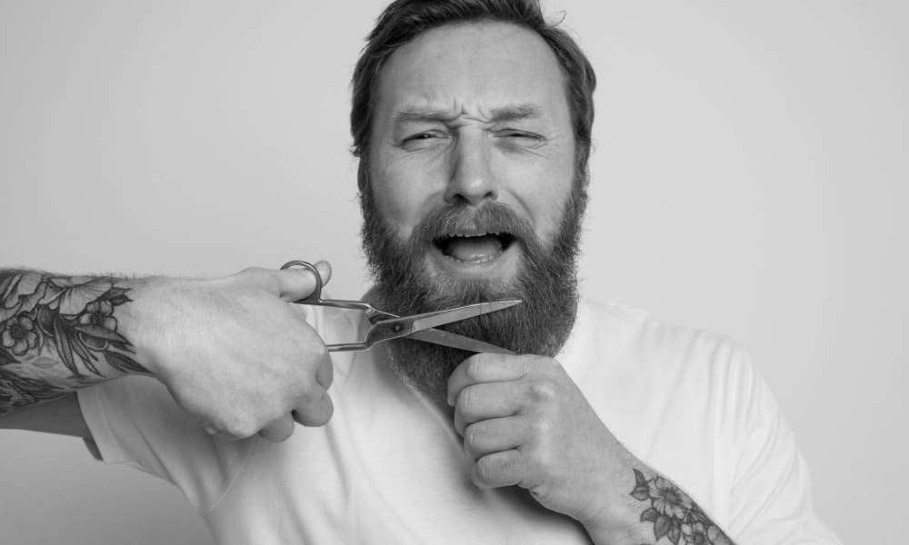 A man with a red beard looking nervous while holding scissors to his beard, mid-trim.