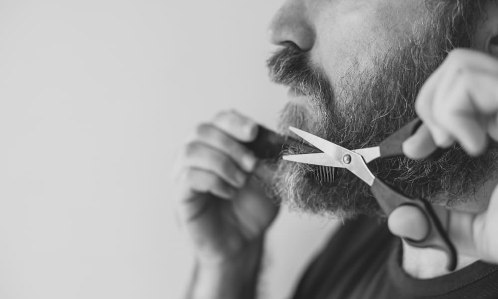 Man trimming his beard with scissors at home, demonstrating how to trim a beard with scissors using a comb for precision.