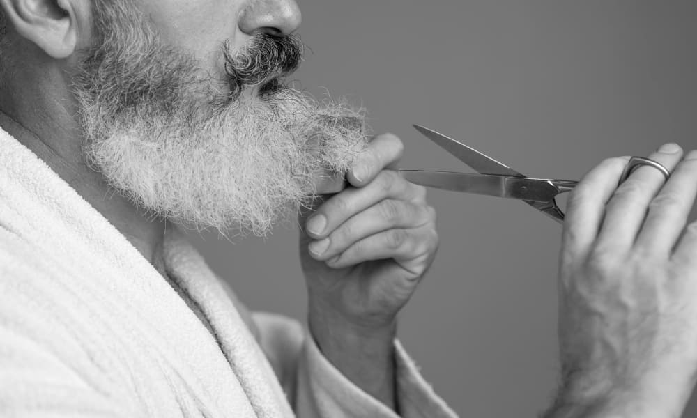 Close-up of an older man grooming his white beard with scissors and a comb.