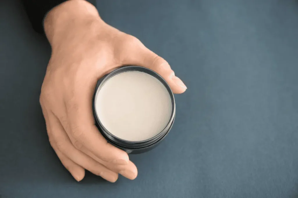 Close-up of men’s hair wax in an open jar held in hand on grey background