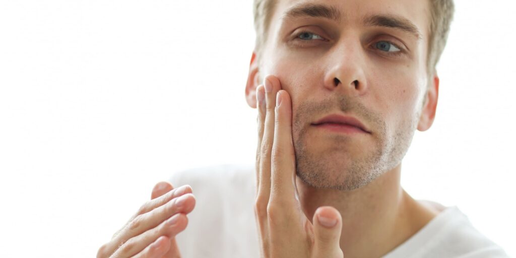 A man applies skincare products to his face. He carefully checks his skin while moisturizing, emphasizing the importance of a daily grooming and skincare routine for healthy, confident skin.