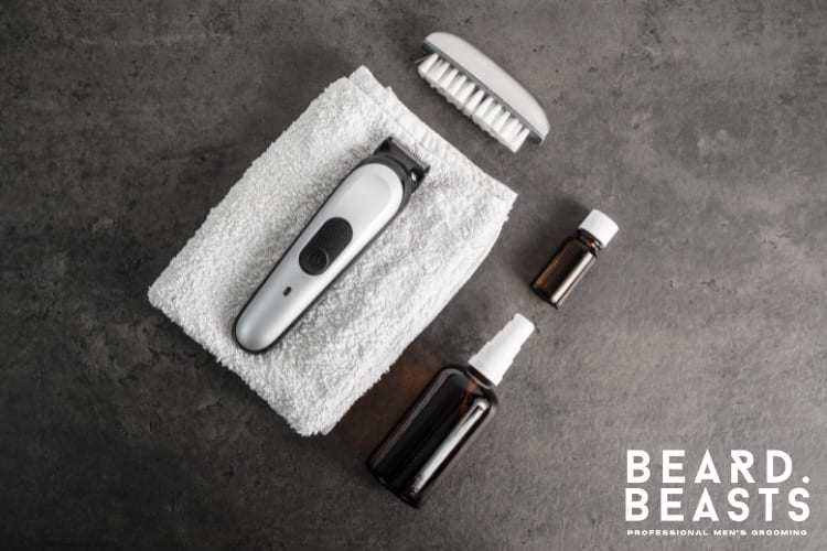 Flat lay of cleaning essentials for grooming tools on a dark surface. The setup includes a white towel, a small cleaning brush, and bottles of cleaning oil and disinfectant spray. The image emphasizes the importance of proper hygiene and maintenance of grooming equipment, perfect for tutorials or care guides.