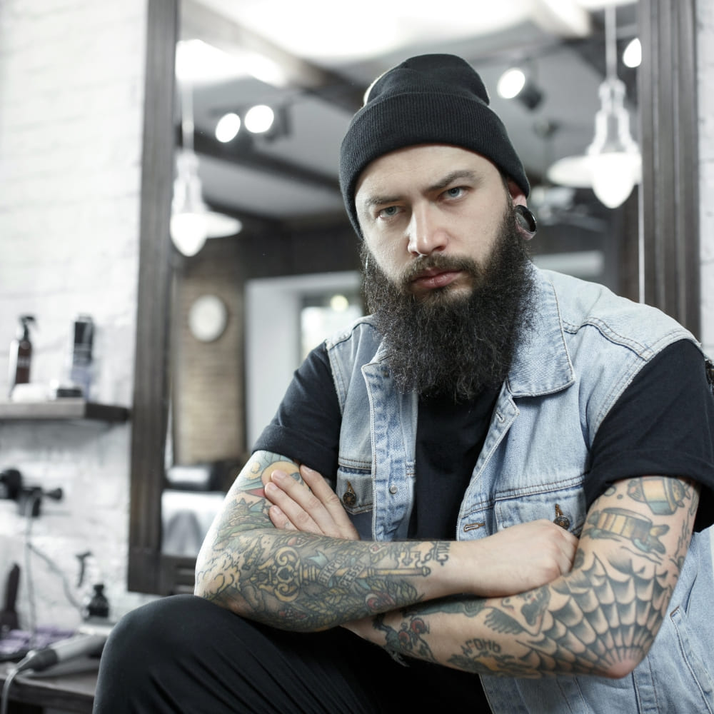 A tattooed man with a long, thick beard and a black beanie, wearing a denim vest over a black shirt, posing confidently in a stylish barbershop. The setting features modern lighting and grooming tools in the background, emphasizing a rugged and trendy aesthetic.