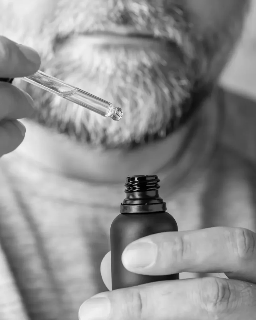 Man applying beard oil with a glass dropper to a grey beard, showing beard oil care for softer facial hair and the skin underneath.