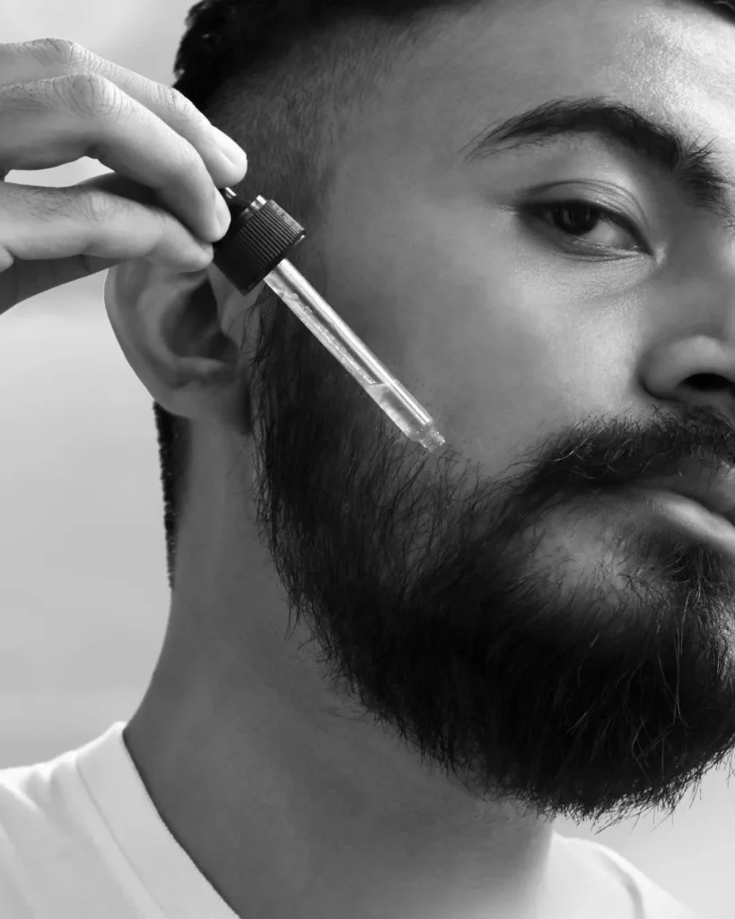 Man applying beard oil with a glass dropper to a full beard, showing beard oil use for softer facial hair and the skin underneath.