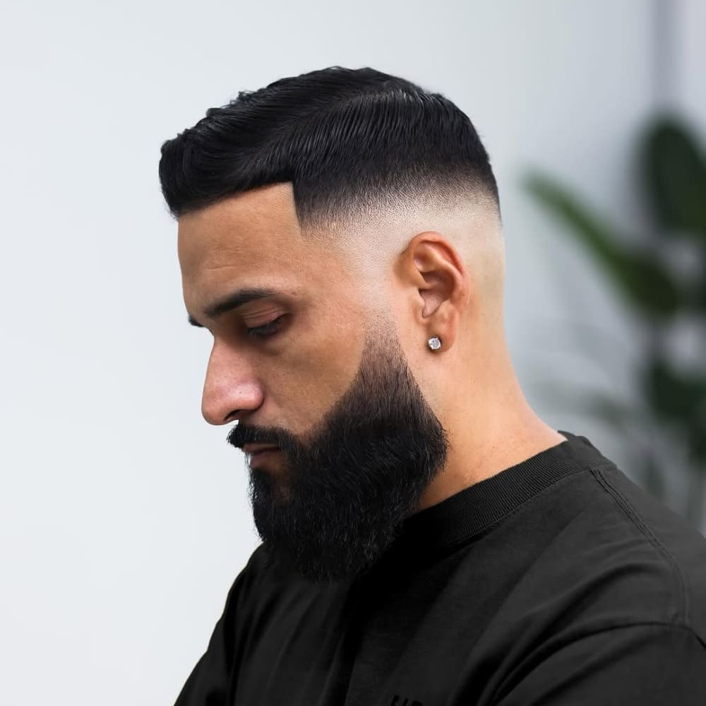 Side profile of a man with a bald fade haircut and a full, well-groomed beard, featuring a seamless fade from the skin at the sides and back to slightly longer, neatly combed hair on top. The bald fade creates a sharp, polished look, emphasizing the contrast between the closely shaved sides and the textured top.