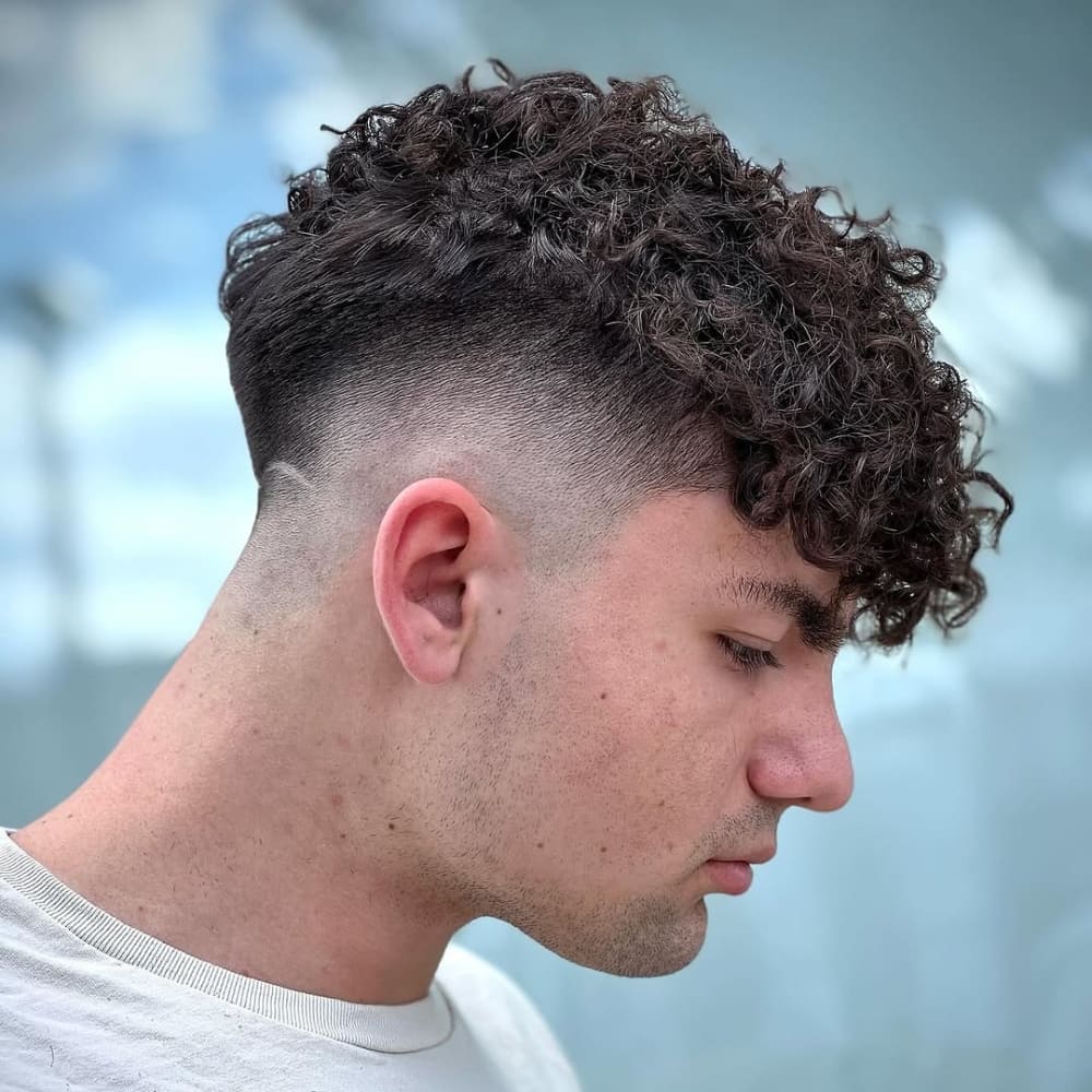 Side profile of a young man with closely cropped sides that transition into thick, curly hair on top. The style emphasizes the natural texture of the curls while maintaining a clean and polished appearance.