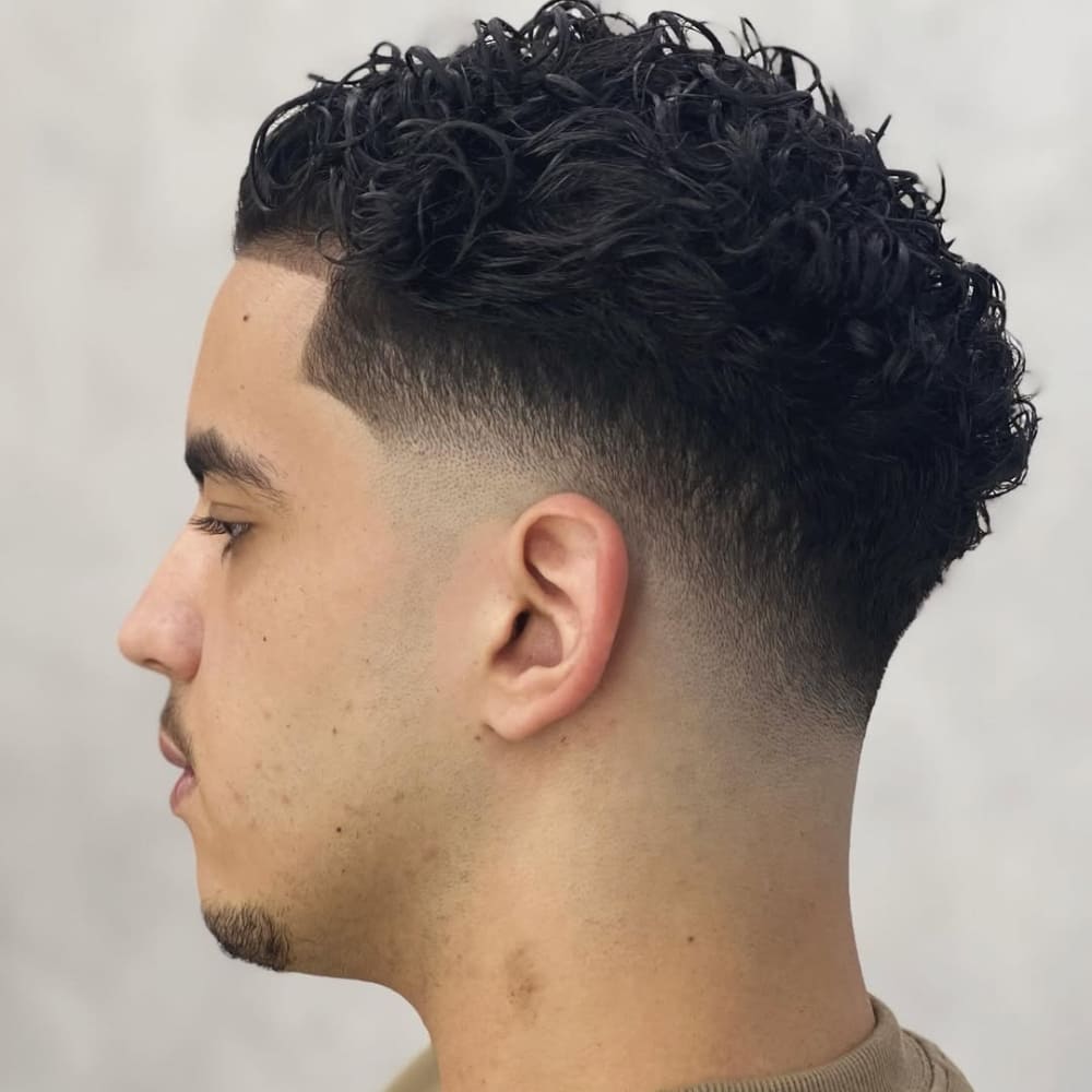 Side profile of a young man with a drop fade haircut, characterized by a fade that curves downward behind the ears, creating a clean and dramatic gradient. The haircut transitions smoothly from skin-level at the sides and back to thick, textured curls on top, offering a modern and stylish appearance.