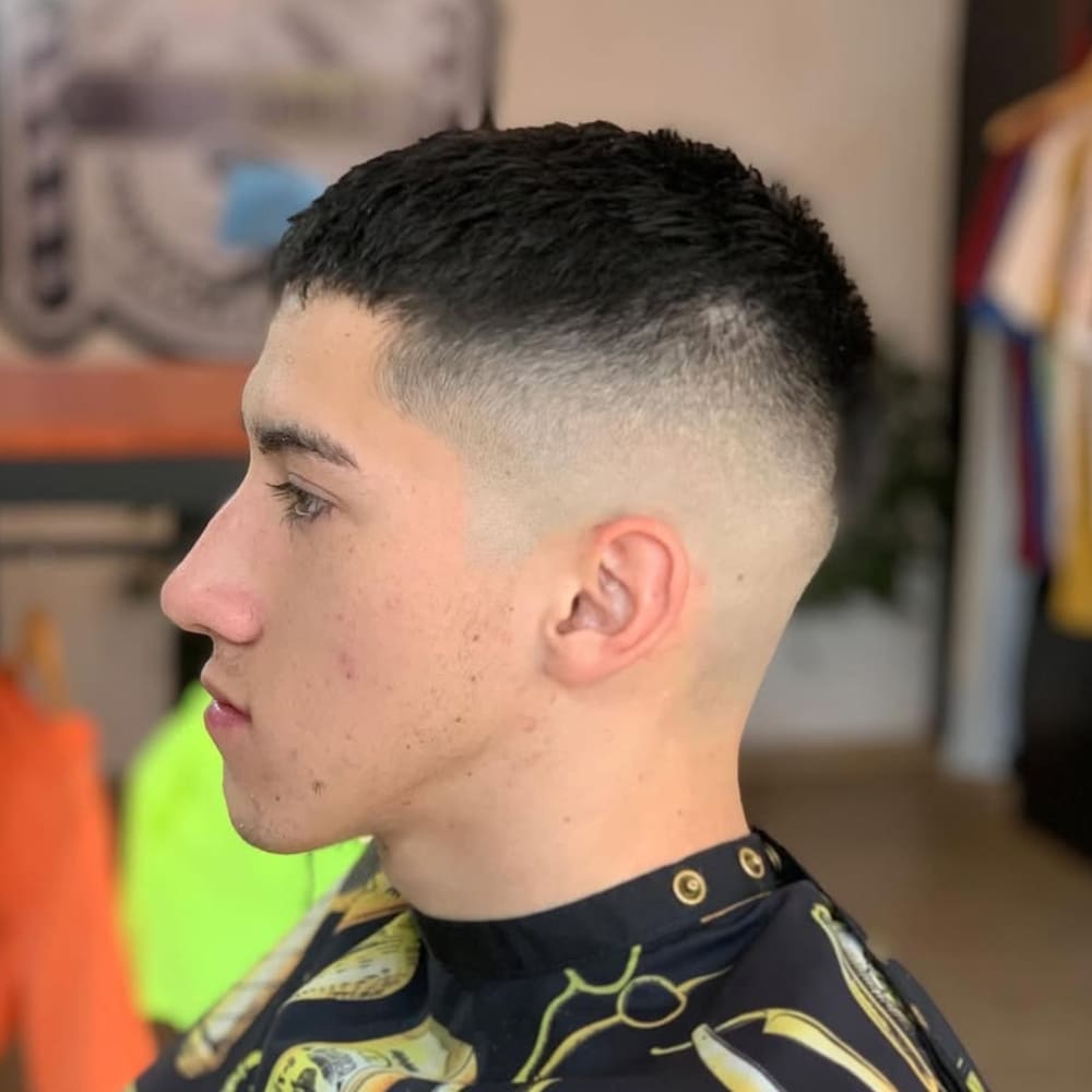 Side profile of a young man with a high fade haircut, featuring a clean transition from very short hair on the sides and back to slightly longer hair on top. A fade haircut is a popular style where the hair tapers gradually, creating a sleek and polished appearance that complements various face shapes and styles.