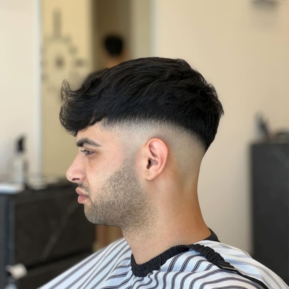 Side profile of a young man with a mid fade haircut, showcasing a smooth gradient from skin-level hair at the sides to longer, textured hair on top. A fade haircut is a versatile style where the hair transitions gradually in length, offering a clean and modern look suitable for various hair textures and face shapes.