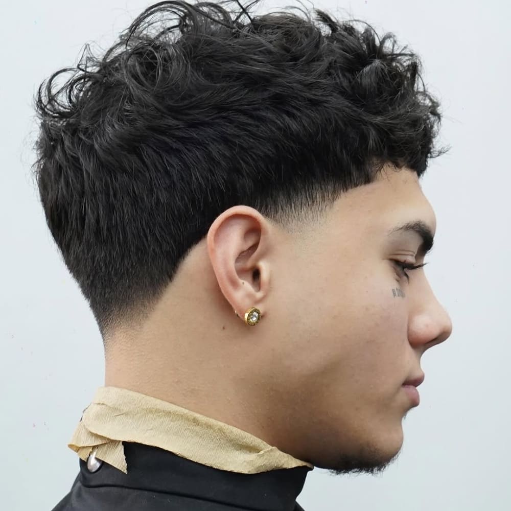 Side profile of a young man with a taper fade haircut, showcasing a gradual fade that blends from the hairline near the ears to longer, textured curls on top. A taper fade is a classic haircut where the hair progressively shortens towards the neckline, providing a clean and balanced appearance while maintaining length and volume on top.