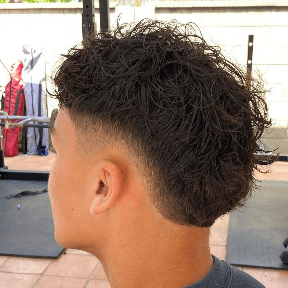 Side profile of a man with a Burst Fade haircut, featuring wavy hair on top that gradually transitions into a fade on the sides and back. The waves are textured and voluminous, adding a natural flow to the hairstyle. The fade starts at the temples, creating a smooth and clean taper that enhances the wavy texture.