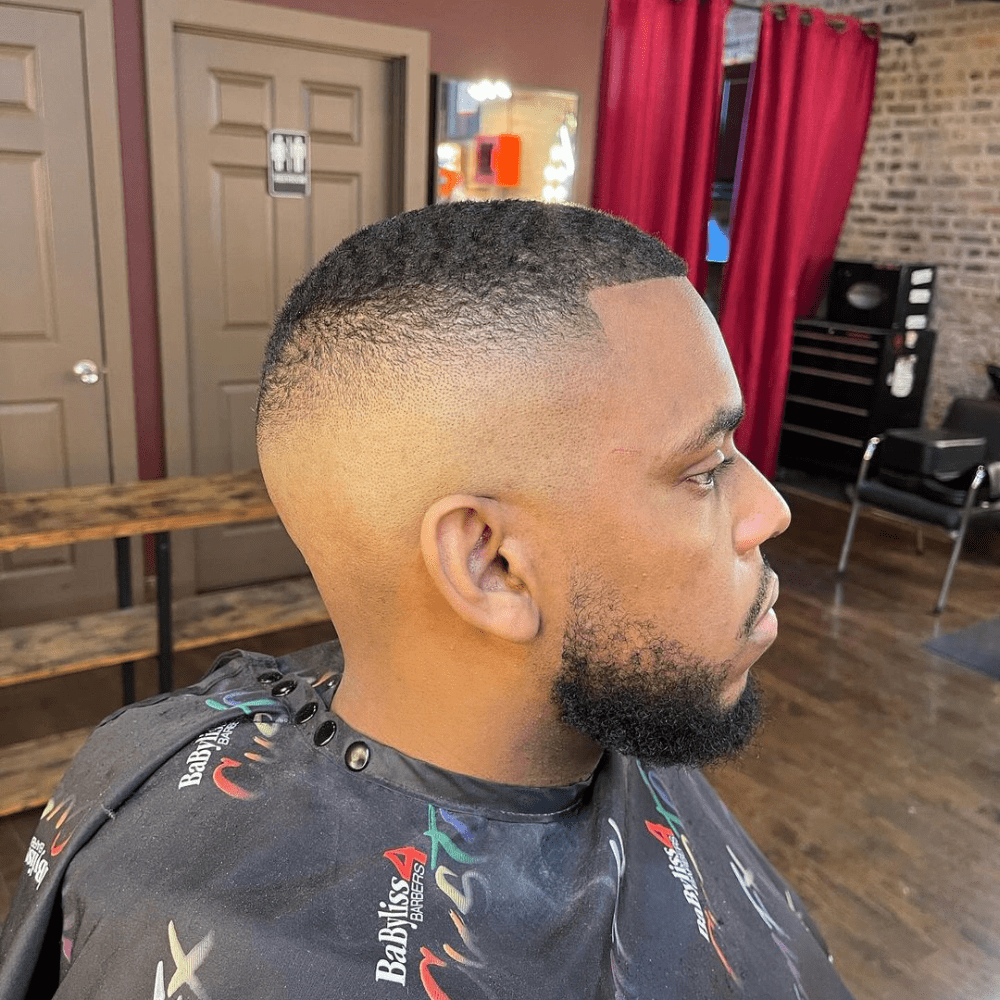 Clean taper with a bald fade on the sides and textured top for a sharp and modern look, paired with a well-groomed beard.
