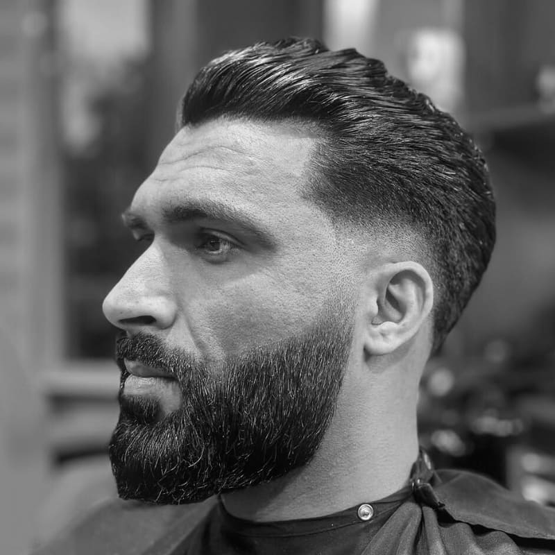 Man in profile showcasing a clean low fade haircut with a voluminous slicked-back top and a neat beard.