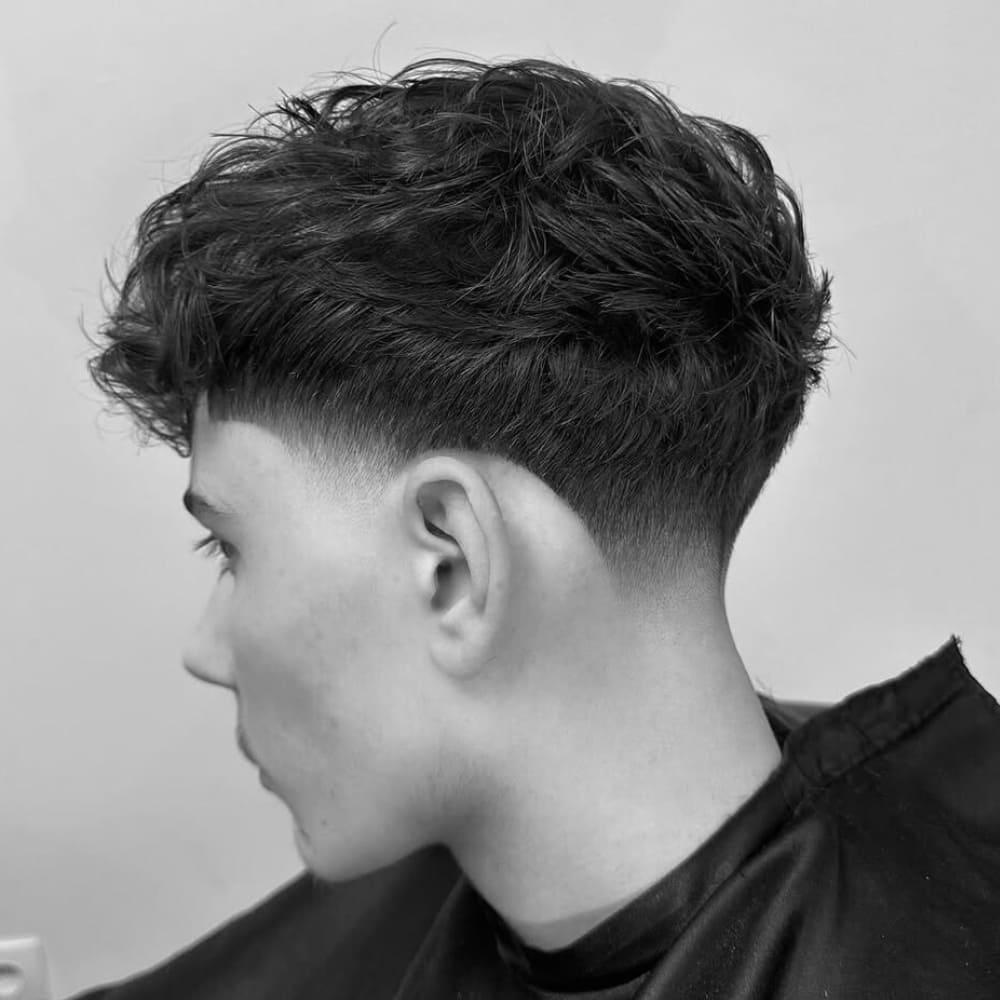 Black and white side profile of a man with a modern taper hairstyle. The haircut features a smooth, gradual fade from longer, textured hair on top to shorter, neatly blended sides. The top hair has a natural, tousled look, giving the style a relaxed yet polished appearance.