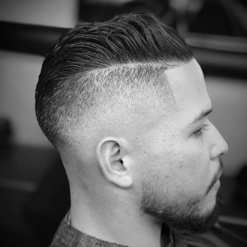 Black and white side profile of a man with a sharp undercut hairstyle. The haircut features closely shaved sides with a clear, defined line separating the longer, slicked-back hair on top. The style is complemented by a neatly trimmed beard, showcasing a modern and clean look