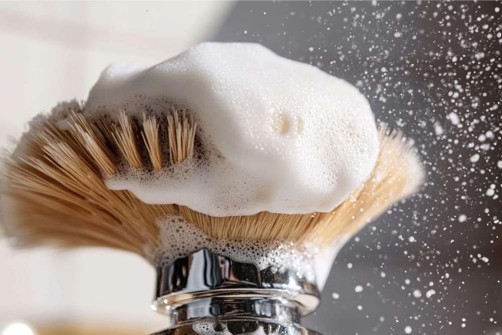 A close-up of a traditional brush with dense natural bristles, covered in rich, foamy lather. Droplets of lather are suspended mid-air, creating a dynamic and refreshing scene. The polished metal base of the brush contrasts beautifully with the light, airy texture of the foam, emphasizing cleanliness and elegance in grooming.