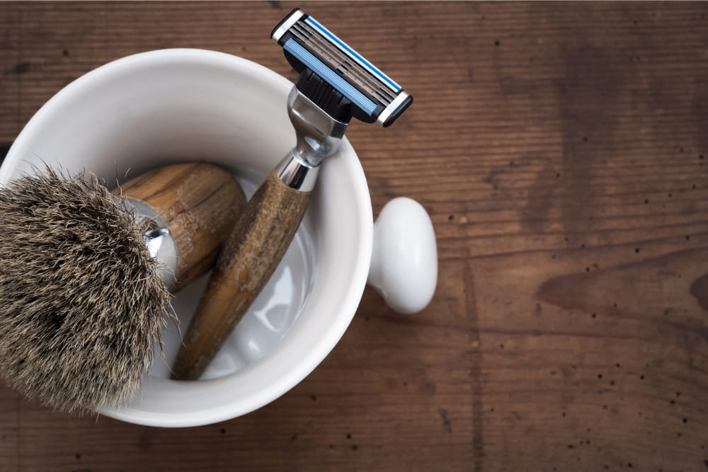 A top-down view of a classic grooming setup featuring a traditional brush with natural bristles and a wooden handle, a modern razor with a sleek design, and a white ceramic bowl. The items are arranged neatly on a rustic wooden surface, showcasing a blend of traditional and contemporary grooming tools in an elegant and minimalistic style.