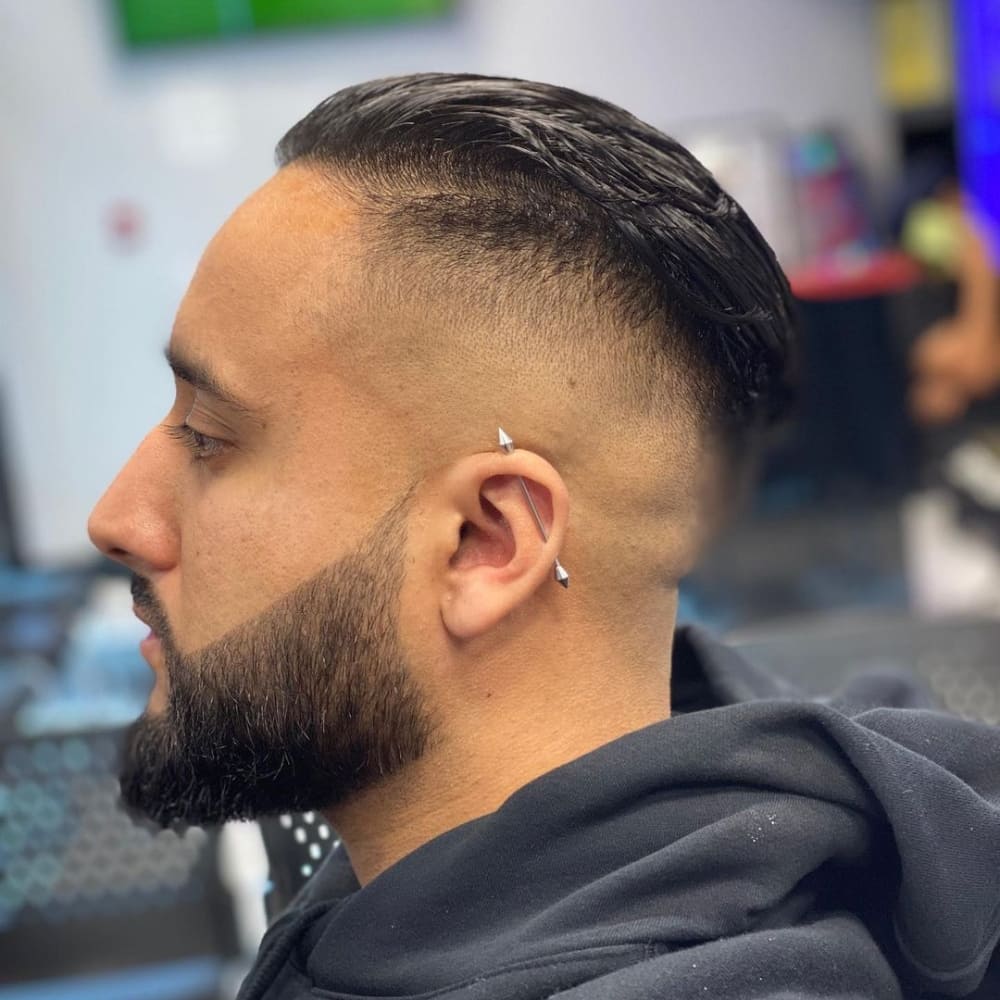 The image shows a person with a slicked-back style. The sides and back of the head are shaved close to the skin, creating a clean high fade. The top hair is left significantly longer and is slicked back smoothly, giving a polished and sleek appearance. The person's beard is neatly trimmed, complementing the sharp lines of the haircut. Additionally, the person has an ear piercing with a barbell earring, adding a modern and edgy touch to the overall look. The combination of the slicked-back top and the high fade creates a stylish and contemporary appearance suitable for various settings, from professional to casual.