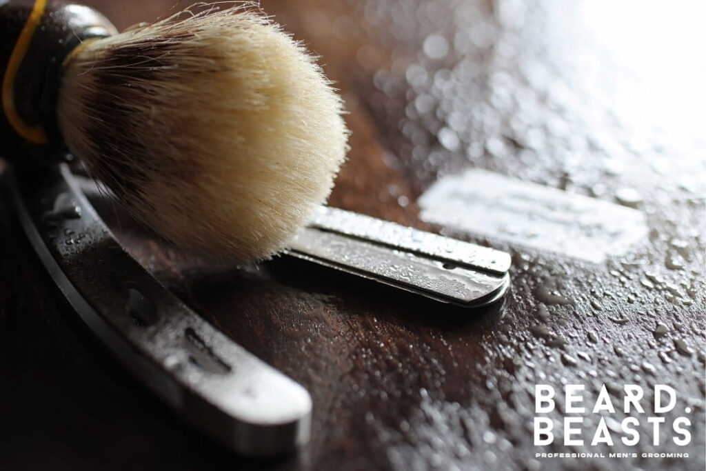 A close-up of a wet shaving brush resting on a wooden surface alongside a razor blade, emphasizing the importance of proper maintenance.