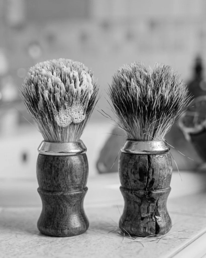 Two shaving brushes showing the difference between soap buildup on a dirty brush and permanent damage from a cracked handle and loose bristles.