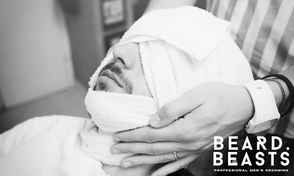 Man receiving a hot towel wrap in a barbershop, relaxing as the steam opens pores and softens facial hair before shaving.