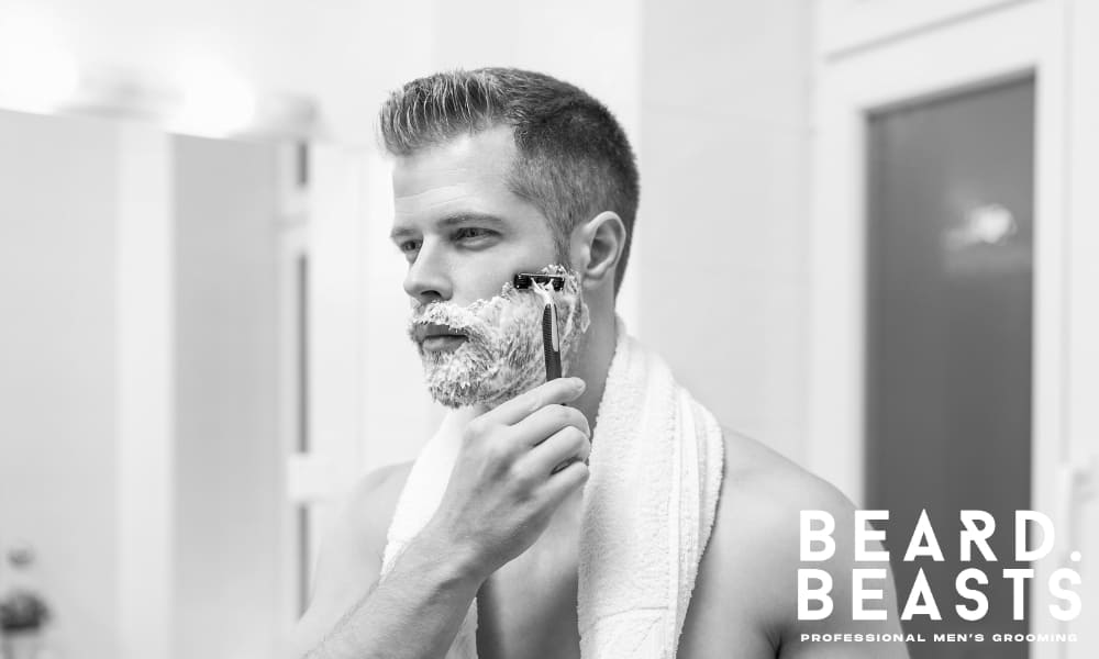 Man applying wet shaving technique in bathroom mirror with full lather and razor, wearing a towel around his neck.