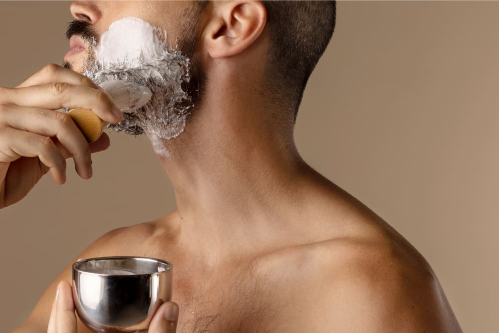 A shirtless man applying lather to his beard using a traditional brush, holding a stainless steel bowl in his other hand. The rich foam covers his facial hair as he prepares for grooming, demonstrating how to use shaving soap effectively with classic tools for a smooth shave. The neutral background emphasizes the simplicity and focus on the grooming ritual.