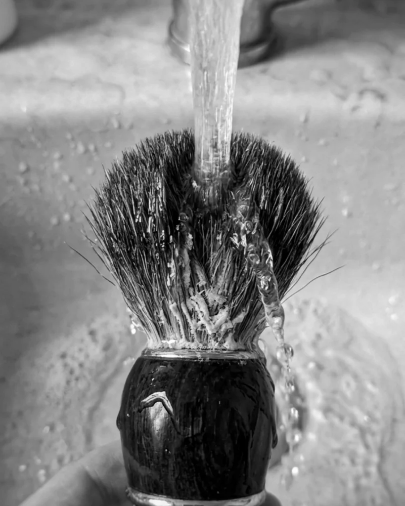 Shaving brush being rinsed under running water in a sink, showing why rinsing alone is not the same as properly cleaning a shaving brush
