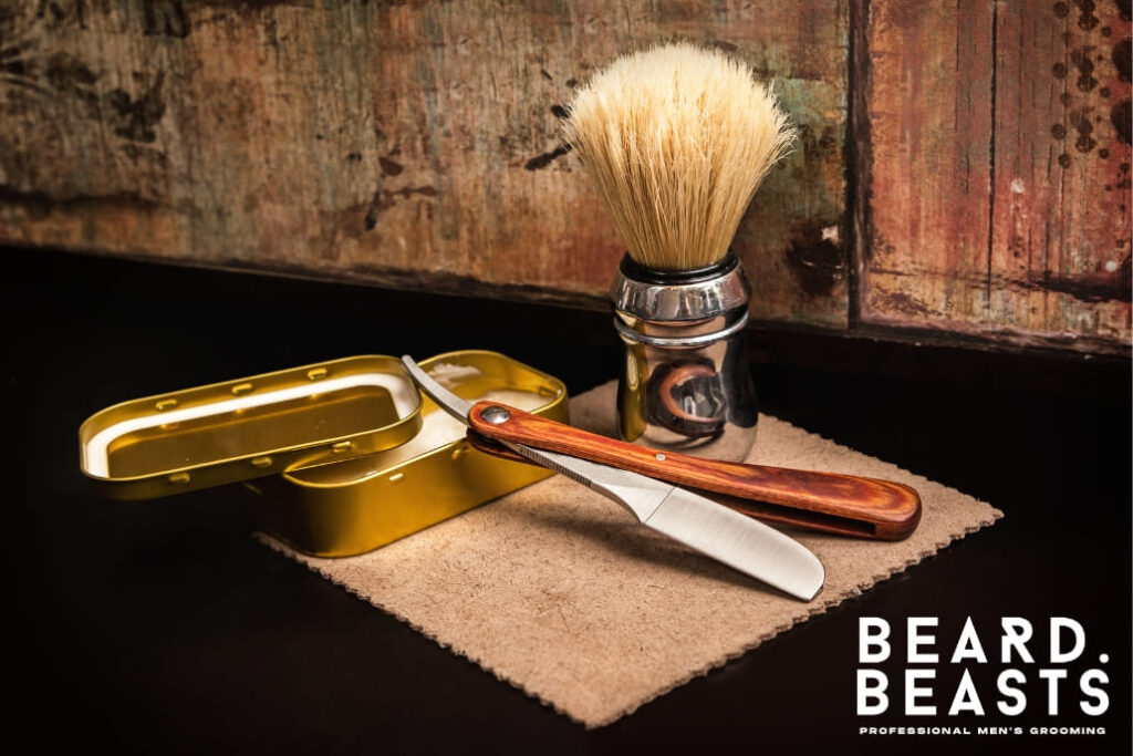 A classic shaving brush with a metal base is displayed alongside a straight razor and a gold tin, set on a rustic wooden surface with a warm, vintage background. Perfect for illustrating traditional grooming essentials and care tips, such as how to clean a shaving brush for optimal performance and hygiene.