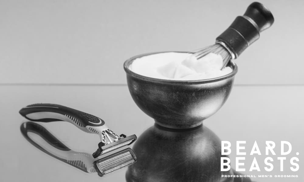 Brush resting in a wooden lather bowl filled with foam beside a modern cartridge razor on a reflective surface.