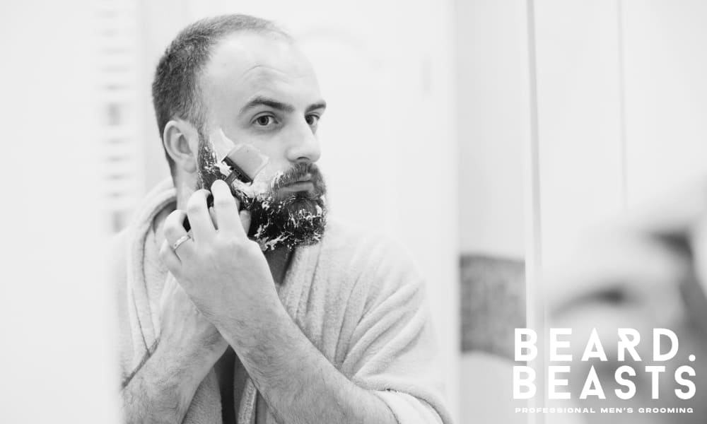 Bearded man carefully shaving in bathroom mirror with lather on face, demonstrating wet shaving technique in robe.