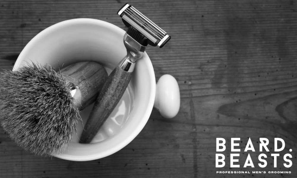 High-angle view of traditional wet shaving tools—wood-handled razor and shaving brush in a white mug on a rustic wooden surface.