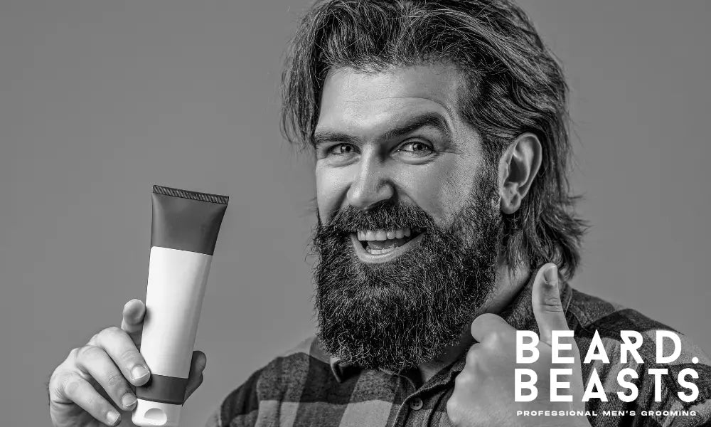 A happy man with a large, well-groomed beard smiling and giving a thumbs-up while holding a tube of beard conditioner.